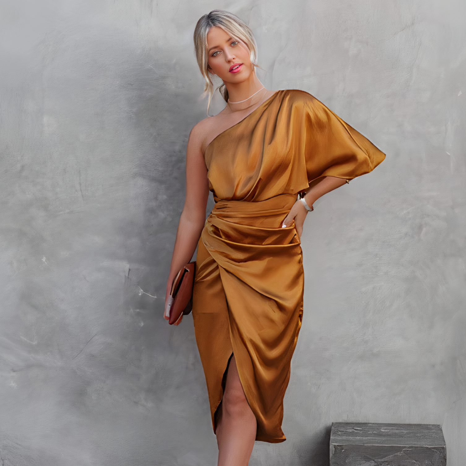 Femme en robe asymétrique dorée satinée, tenant une pochette marron, posant devant un mur gris texturé. Mode élégante, tenue de soirée chic.