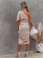 Femme en robe asymétrique beige, sac blanc à la main, posant devant un mur texturé avec vase et herbes pampas. Mode élégante et chic.