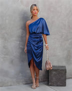 Femme en robe bleue asymétrique élégante, tenant un sac à main rose, posant contre un mur gris. Mode féminine chic, style moderne.
