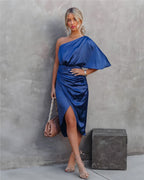 Femme en robe bleue asymétrique élégante, posant contre un mur gris, tenant un sac à main beige. Mode chic, style moderne, tenue soirée.