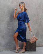 Femme en robe bleue élégante, posant contre un mur gris, tenant un sac à main beige. Mode chic, style soirée, tendance 2023.