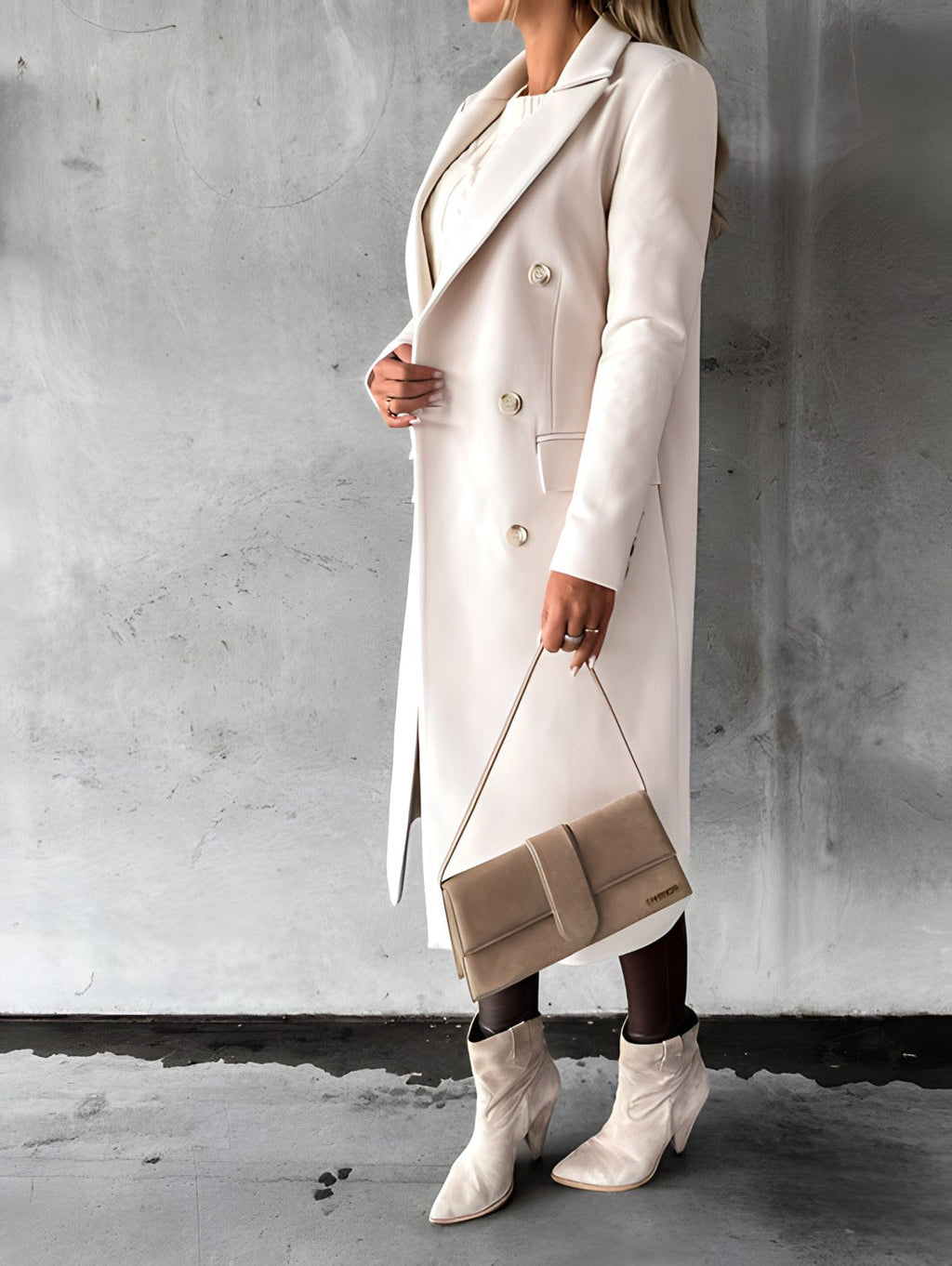 Femme en manteau beige élégant, tenant un sac à main assorti, portant des bottes blanches, posant devant un mur gris. Mode automne chic.