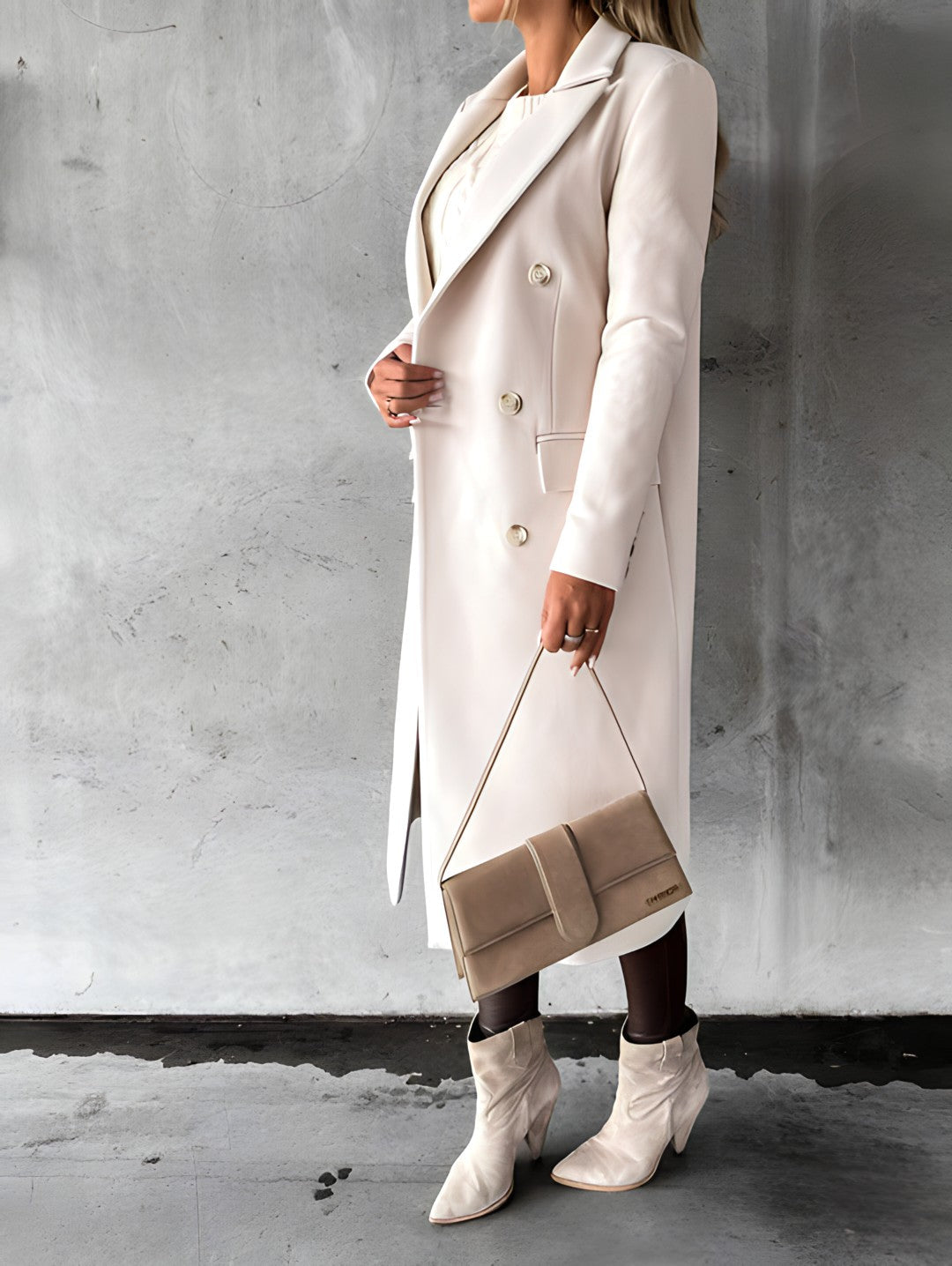 Femme en manteau beige élégant, tenant un sac à main assorti, portant des bottes blanches, posant devant un mur gris. Mode automne chic.