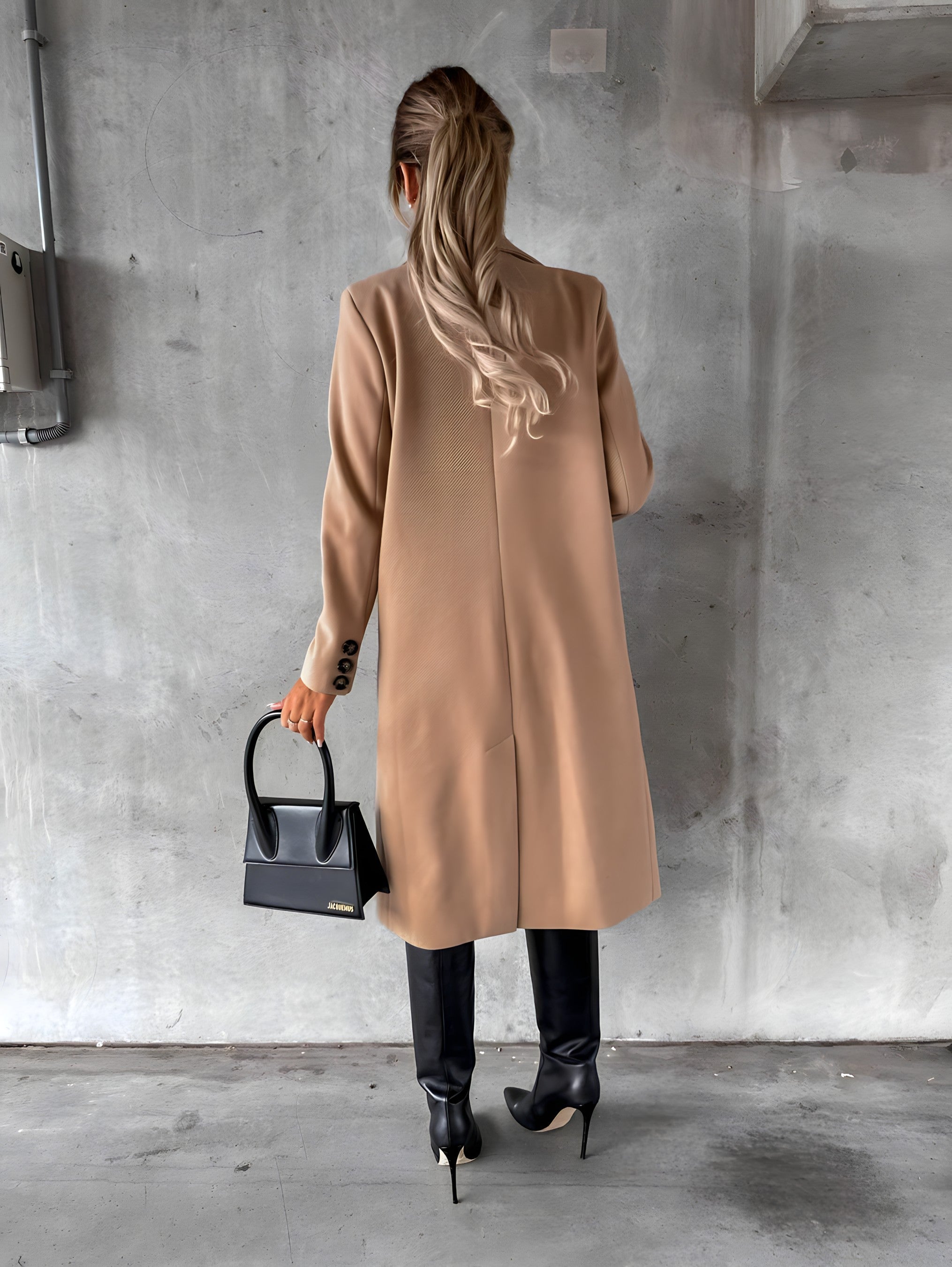 Femme de dos portant un manteau beige, bottes noires et tenant un sac à main noir, posant contre un mur en béton. Mode élégante automne-hiver.