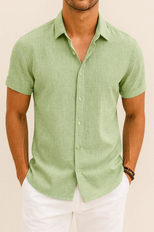 Homme portant une chemise en lin vert clair à manches courtes, col classique, boutons blancs, associée à un pantalon blanc. Mode estivale élégante.