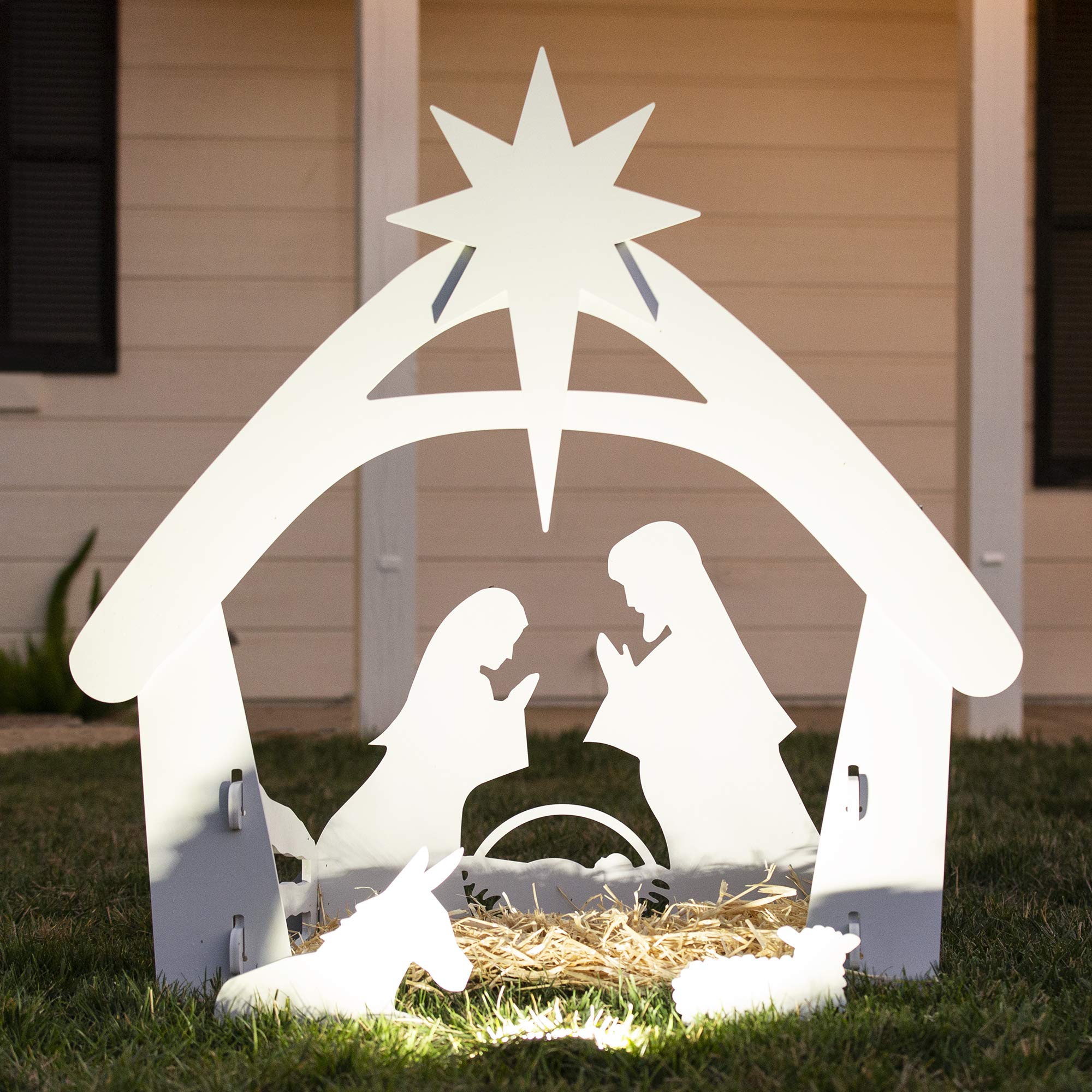 Crèche de Noël en bois blanc avec étoile, représentant Marie, Joseph et Jésus, éclairée dans un jardin. Décoration festive extérieure.