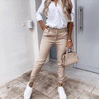 Femme en chemise blanche et pantalon beige, tenant un sac à main beige, portant des baskets blanches, posant devant une porte moderne.
