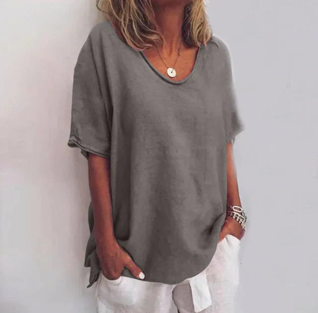 Femme portant un t-shirt gris ample, col rond, manches courtes, avec un collier pendentif et un bracelet, style décontracté estival.