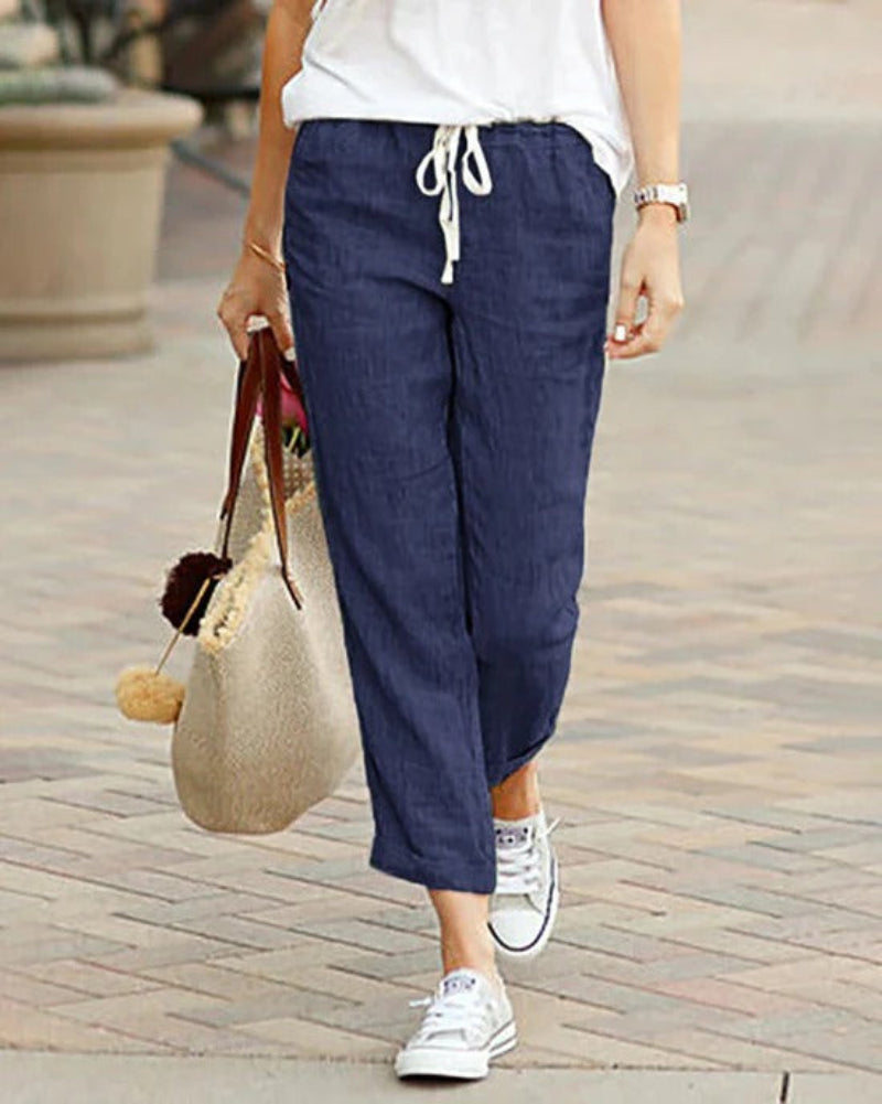 Femme portant un pantalon en lin bleu, t-shirt blanc, baskets blanches, tenant un sac en paille avec pompons, marchant sur un trottoir.