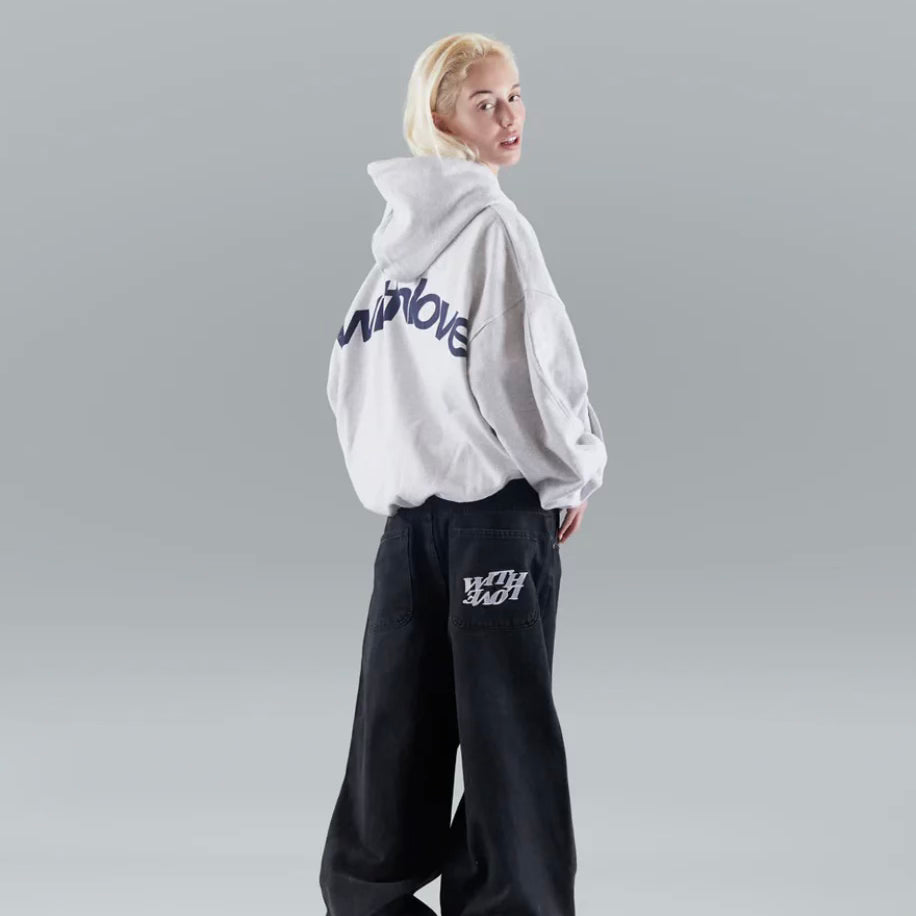 Jeune femme blonde portant un sweat à capuche gris avec inscription, pantalon large noir, pose de dos. Mode urbaine, style décontracté, tendance streetwear.