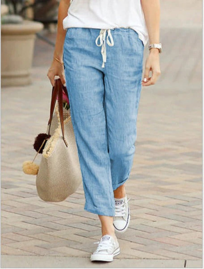 Femme portant un pantalon en lin bleu clair, t-shirt blanc, baskets blanches, sac en paille avec pompons, marchant sur un trottoir pavé.