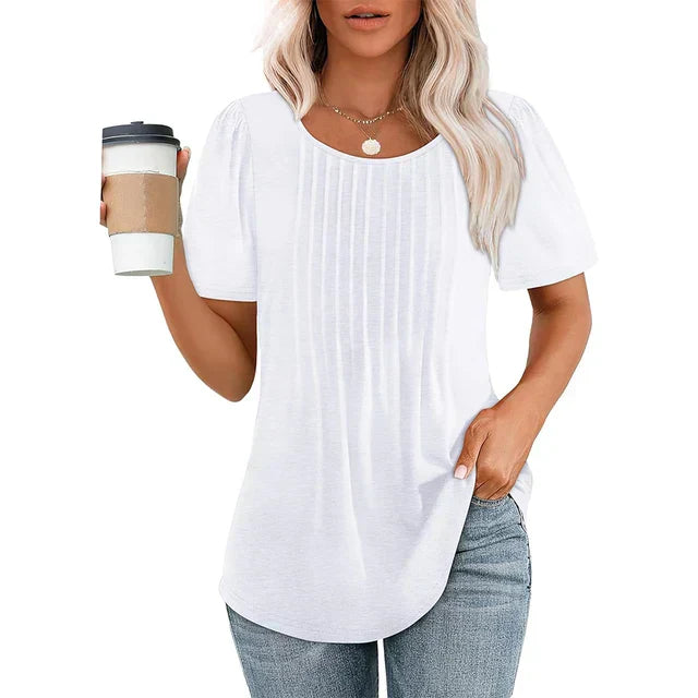 Femme portant un t-shirt blanc plissé, tenant un gobelet de café, cheveux blonds ondulés, collier doré, jeans décontracté. Mode féminine décontractée.