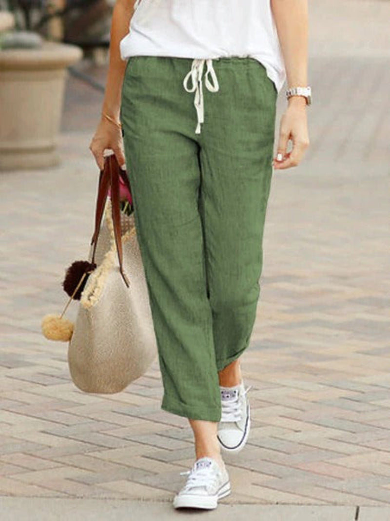 Femme portant un pantalon vert en lin, baskets blanches, et sac beige avec pompons, marchant sur un trottoir. Mode décontractée, style estival.