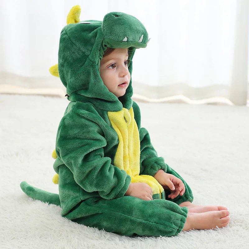 Enfant en costume de dinosaure vert, assis sur un tapis blanc. Pyjama en peluche avec capuche et queue, idéal pour déguisement ou pyjama enfant.