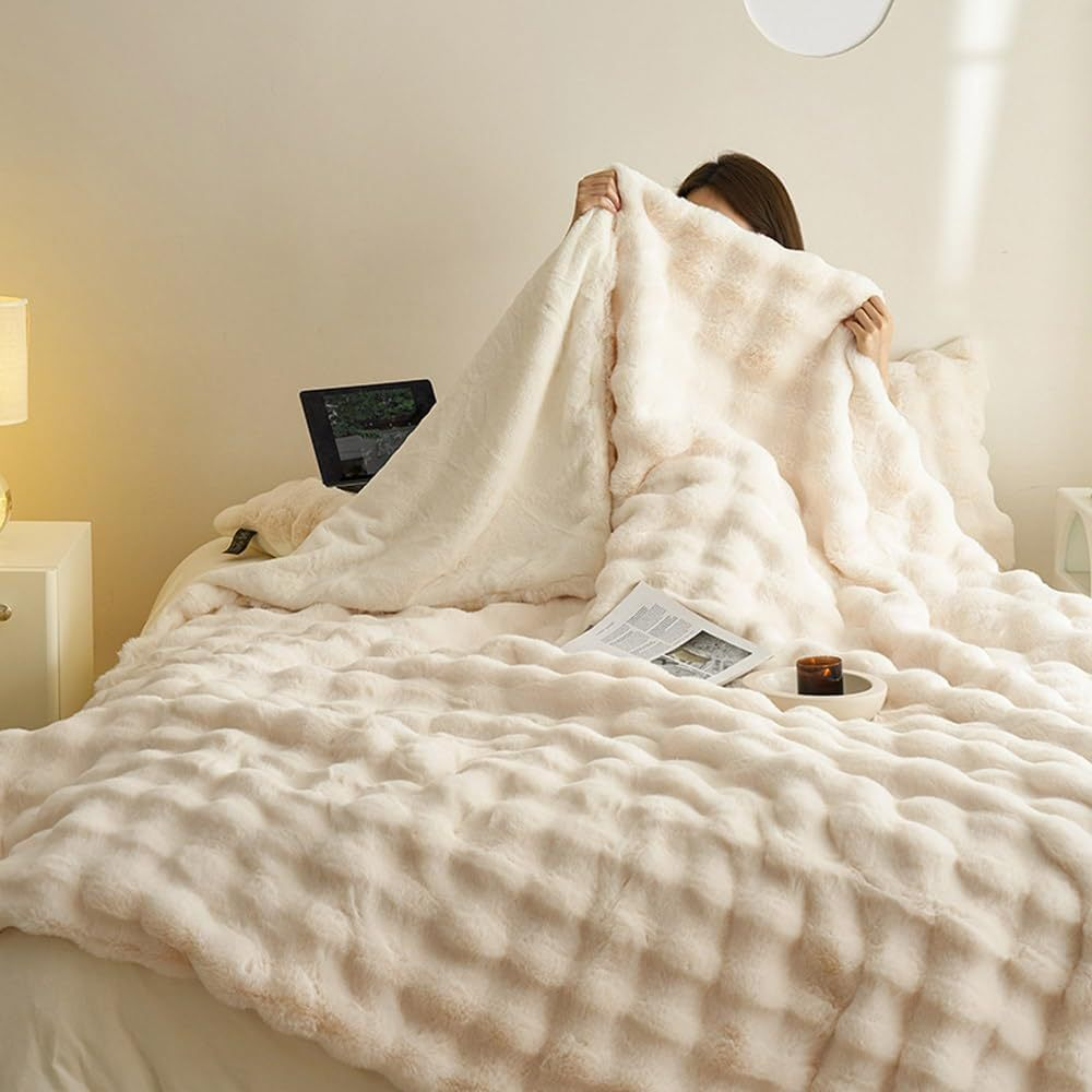 Couverture en fausse fourrure beige sur un lit, femme cachée dessous, ambiance cosy avec lumière douce, livre et bougie sur la table de chevet.