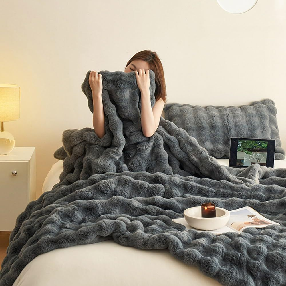 Femme enveloppée dans une couverture en fausse fourrure grise sur un lit, avec une tablette et une bougie allumée sur la table de chevet. Ambiance cosy.