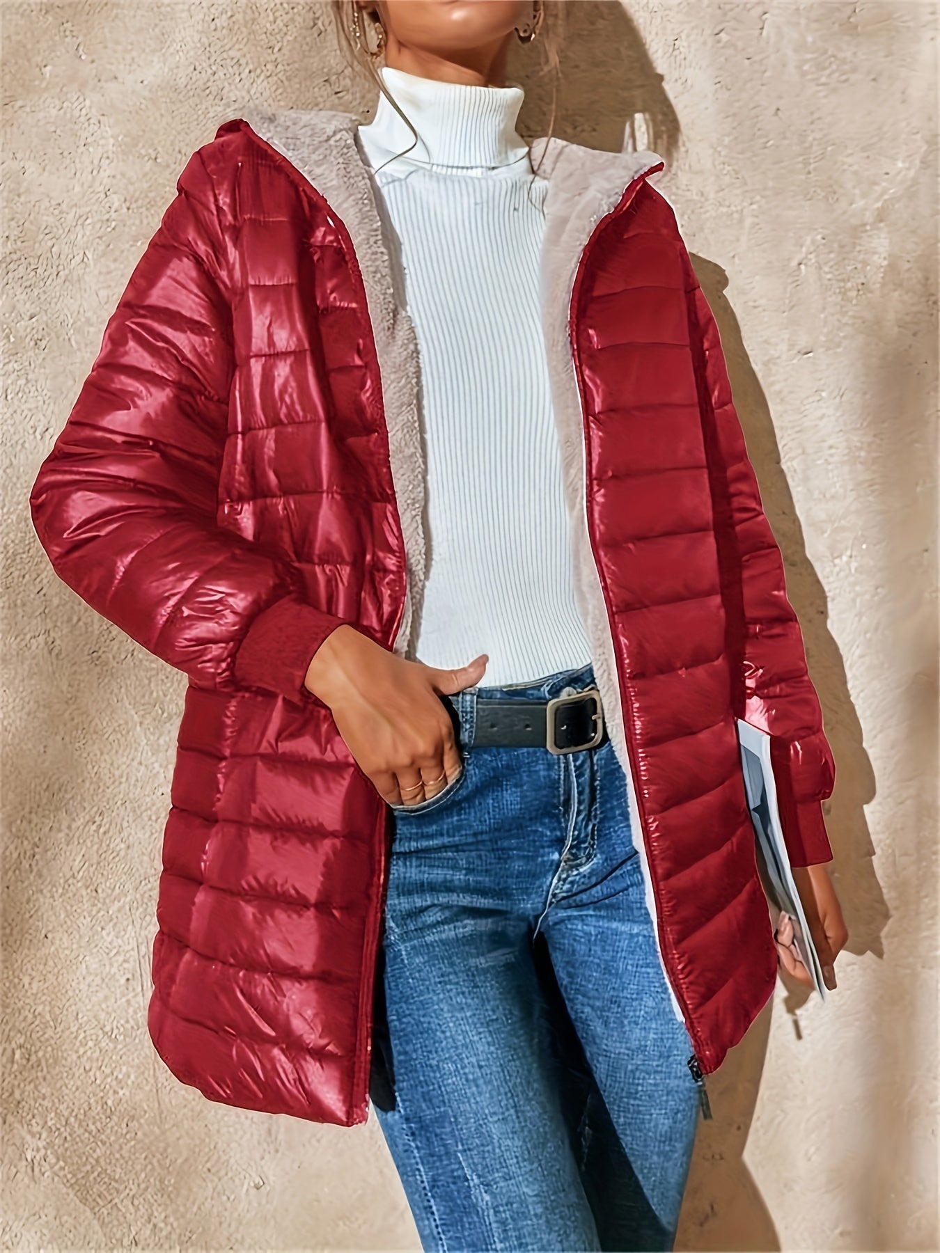 Femme portant une doudoune rouge matelassée, pull blanc à col roulé, jean bleu, tenant un livre. Mode automne-hiver, style décontracté.