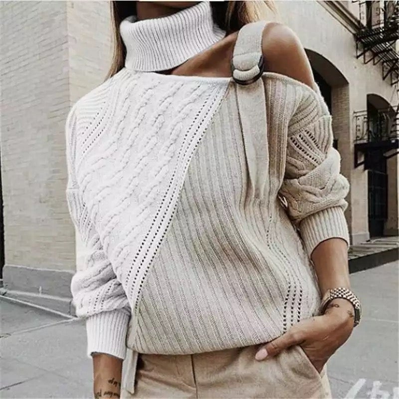 Femme portant un pull asymétrique beige et blanc à col roulé, style urbain chic, manches longues, rue pavée en arrière-plan.