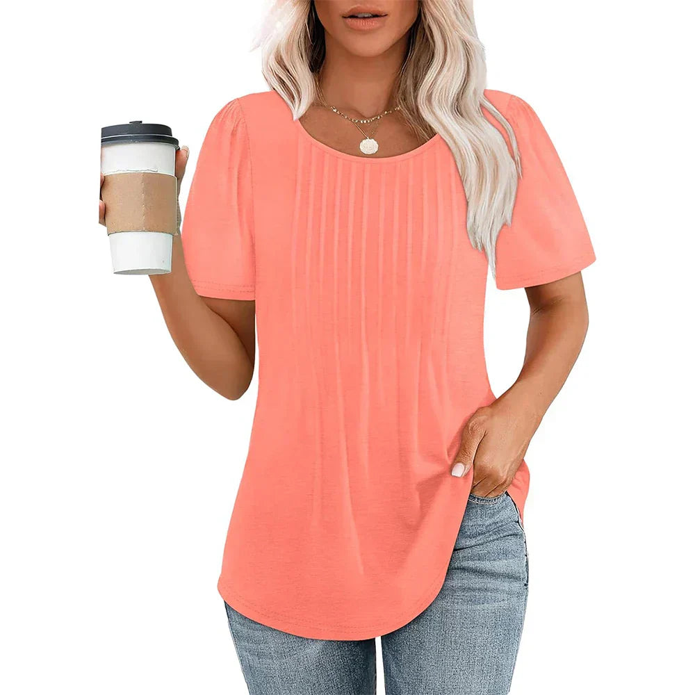 Femme portant un t-shirt corail à manches courtes, tenant un gobelet de café, cheveux blonds ondulés, collier doré, jean décontracté.