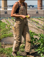 Femme en salopette marron travaillant dans un jardin urbain sur un toit, tenant un légume. Agriculture urbaine, culture durable, potager en ville.