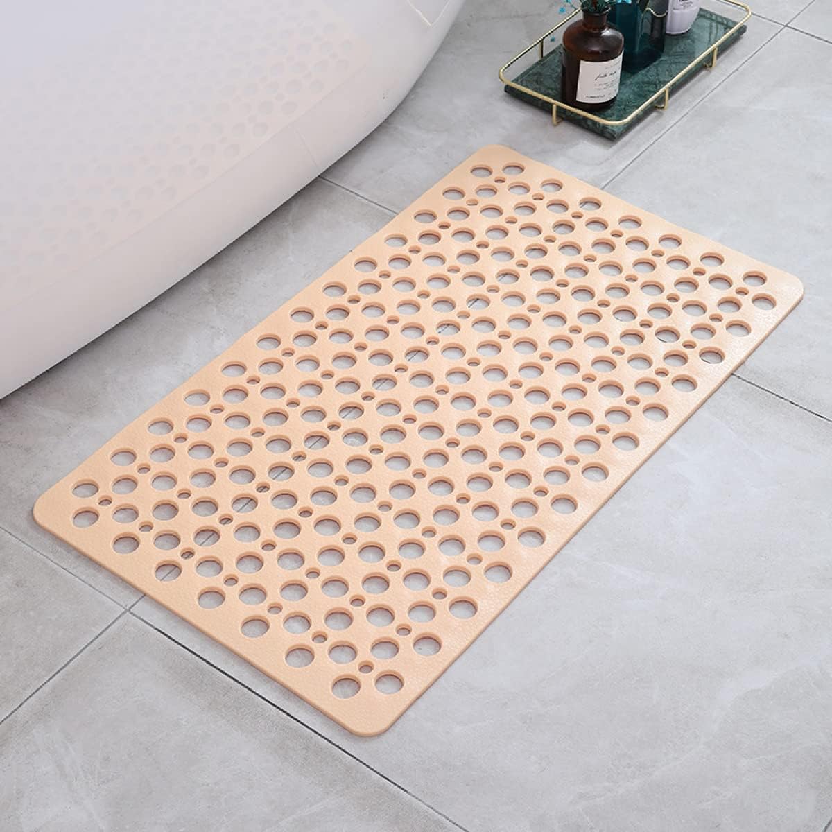 Tapis de bain antidérapant beige avec trous, posé sur carrelage gris, à côté d'une baignoire et d'un plateau avec bouteilles. Accessoire salle de bain.