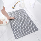 Tapis de bain antidérapant gris avec trous, sur sol de salle de bain en marbre blanc, pied nu et douchette dorée à proximité.