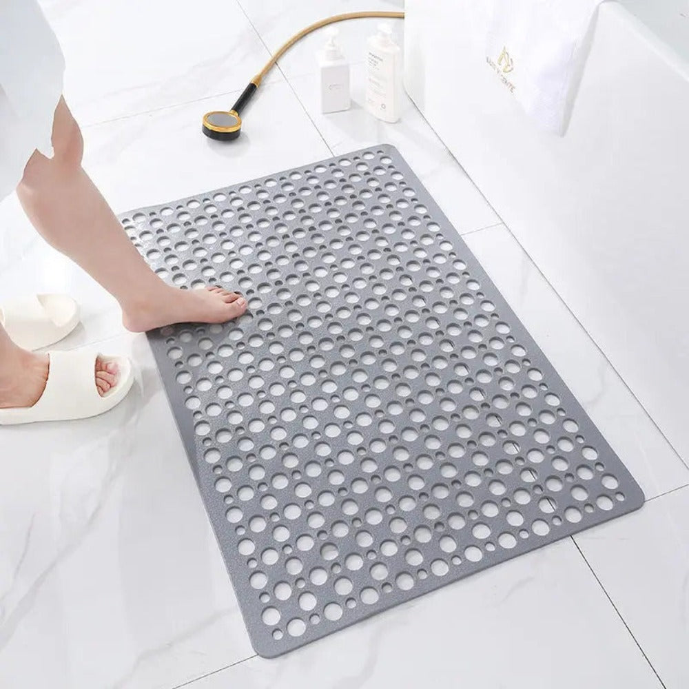 Tapis de bain antidérapant gris avec trous, sur sol de salle de bain en marbre blanc, pied nu et douchette dorée à proximité.