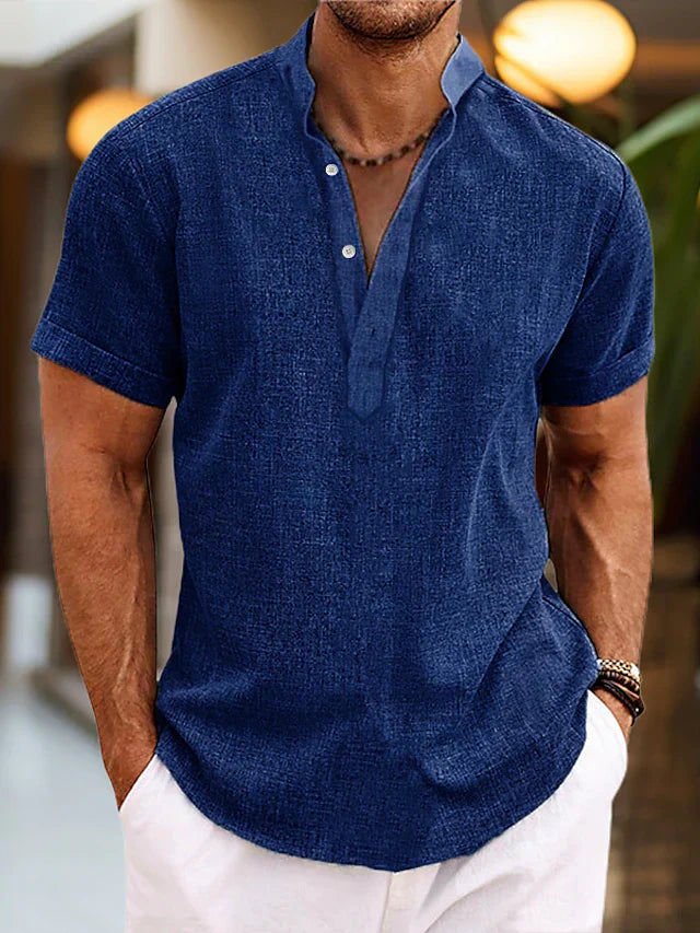 Homme portant une chemise en lin bleu marine à col mao, manches courtes, avec un collier en perles, dans un cadre intérieur moderne.
