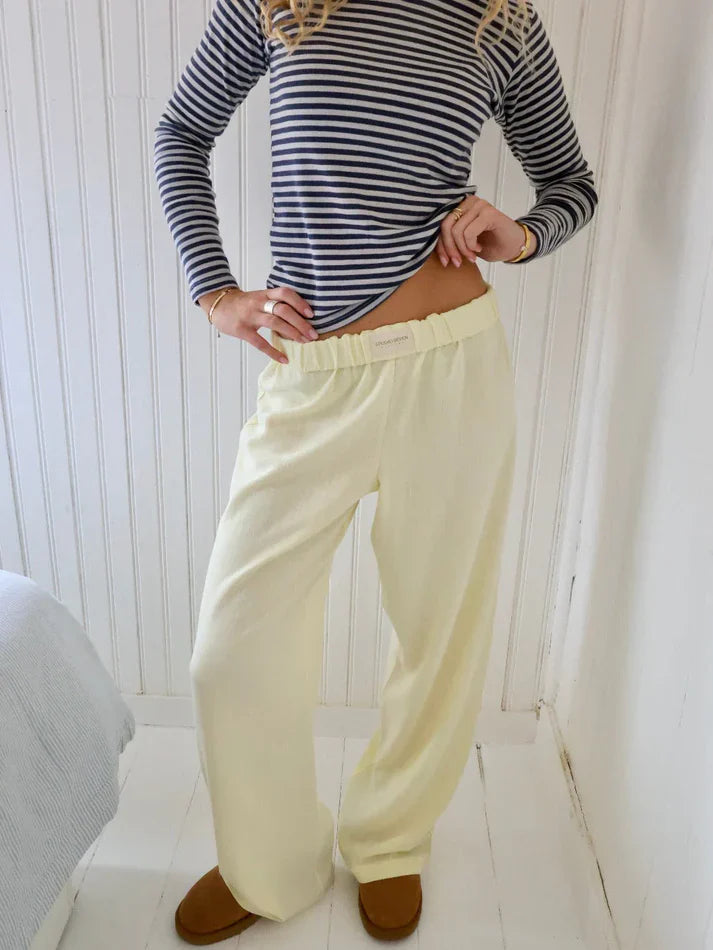 Femme en pull rayé bleu et blanc, pantalon ample jaune clair, posant dans une chambre lumineuse. Mode décontractée, style confortable, intérieur moderne.