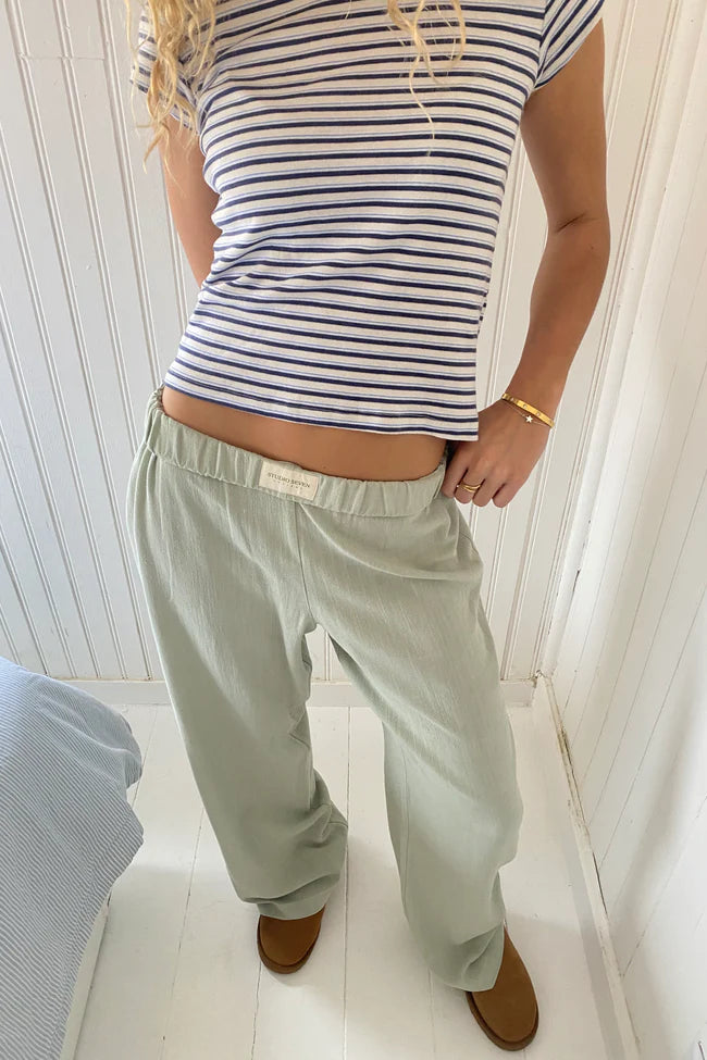 Femme en t-shirt rayé bleu et blanc, pantalon ample vert clair, posant dans une chambre lumineuse. Mode décontractée, style estival, intérieur moderne.