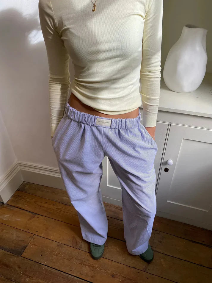 Femme portant un pull crème ajusté et un pantalon ample bleu clair, debout sur un parquet en bois, avec un vase blanc en arrière-plan.