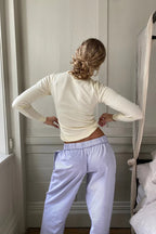 Femme de dos portant un pull beige et un pantalon de pyjama bleu clair, posant dans une chambre lumineuse près d'une fenêtre.