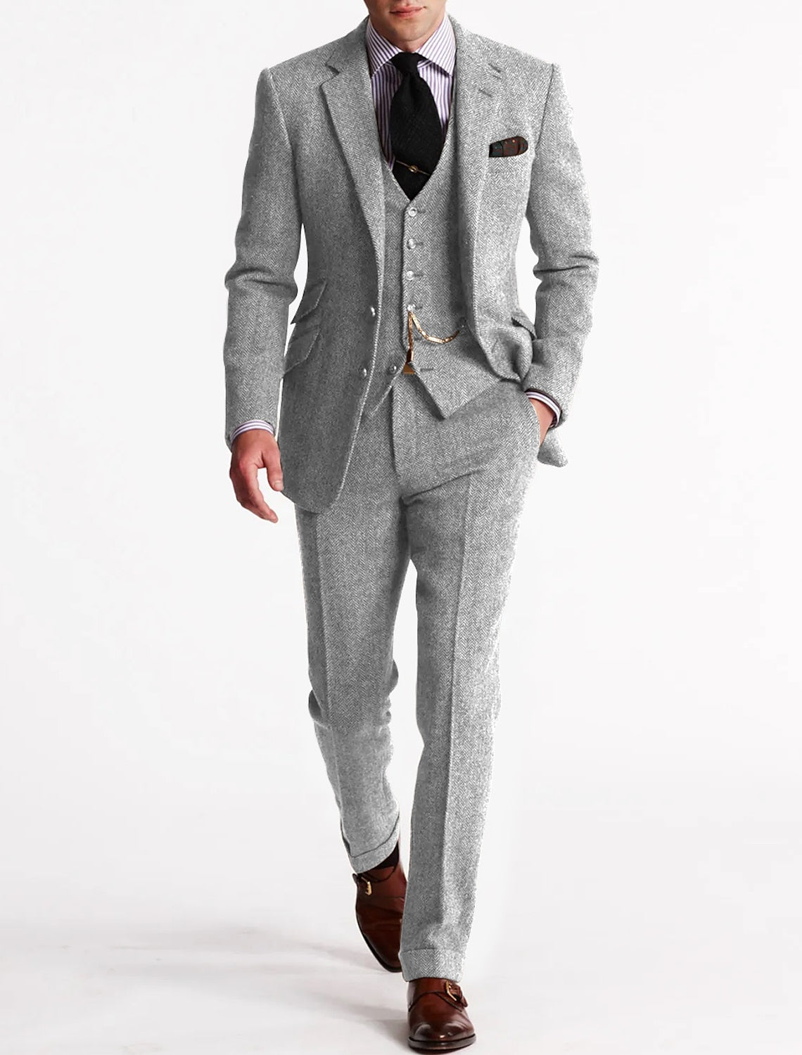 Homme en costume gris élégant, cravate noire, chemise rayée, gilet assorti, chaussures marron, posant sur fond blanc. Mode masculine chic.