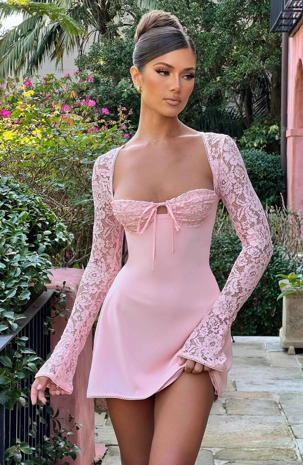 Femme en robe rose élégante avec manches en dentelle, posant dans un jardin fleuri. Mode féminine, style chic, tenue de soirée glamour.
