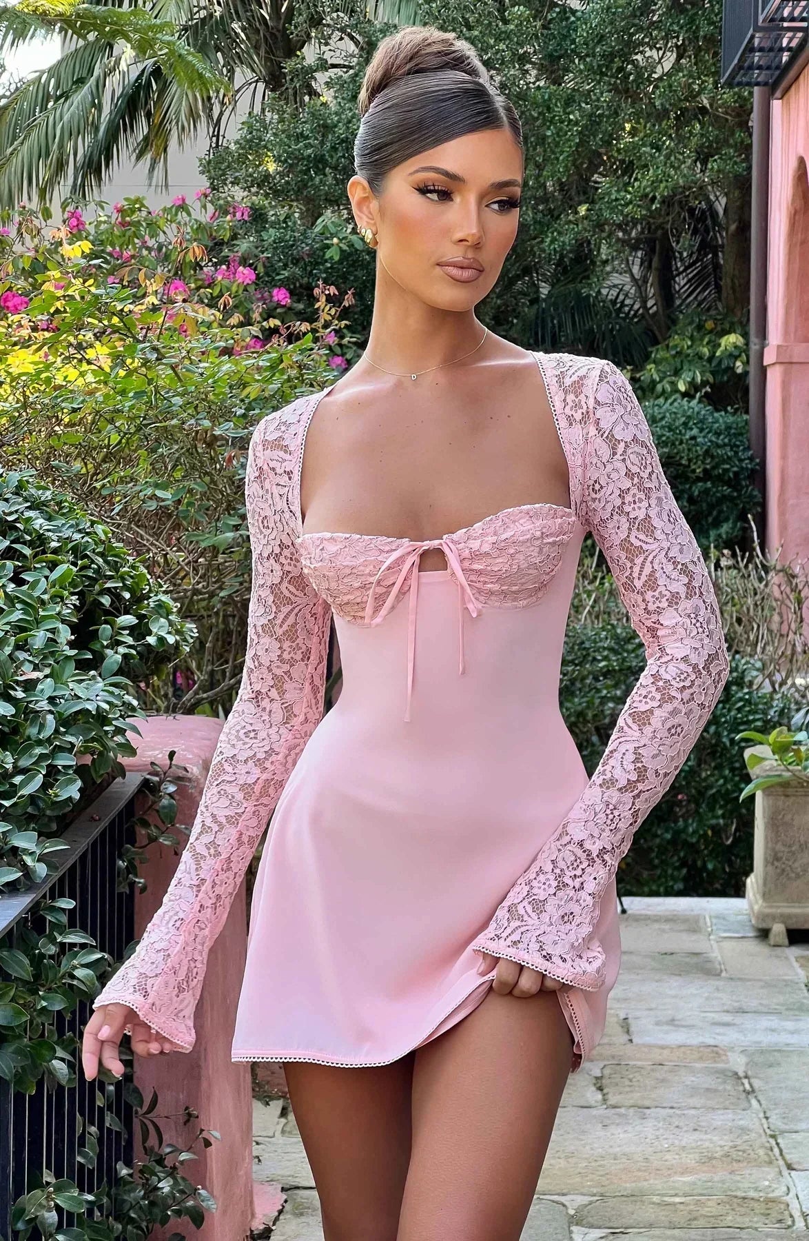Femme en robe rose élégante avec manches en dentelle, posant dans un jardin fleuri. Mode féminine, style chic, tenue de soirée glamour.