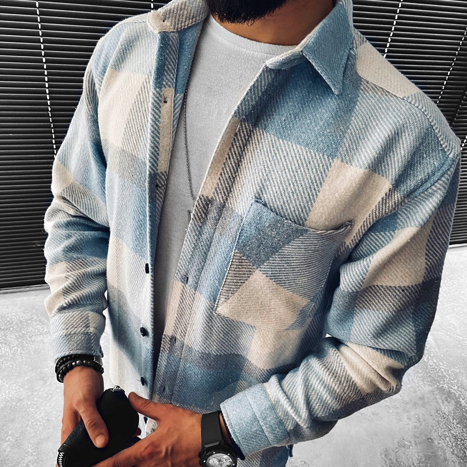 Homme portant une chemise à carreaux bleue et blanche, manches longues, sur un t-shirt blanc, devant des stores noirs. Mode masculine décontractée.