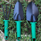 Trois outils de jardinage avec manches verts sur herbe : une fourchette à main et deux pelles à main noires. Accessoires essentiels pour jardin.
