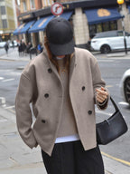 Femme portant un manteau beige à double boutonnage, casquette noire, sac à main noir, marchant en ville. Mode automne, style urbain chic.