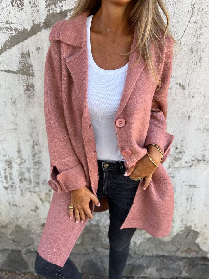 Femme portant un manteau rose élégant, t-shirt blanc et jeans noirs, posant devant un mur texturé. Mode automne, style décontracté chic.
