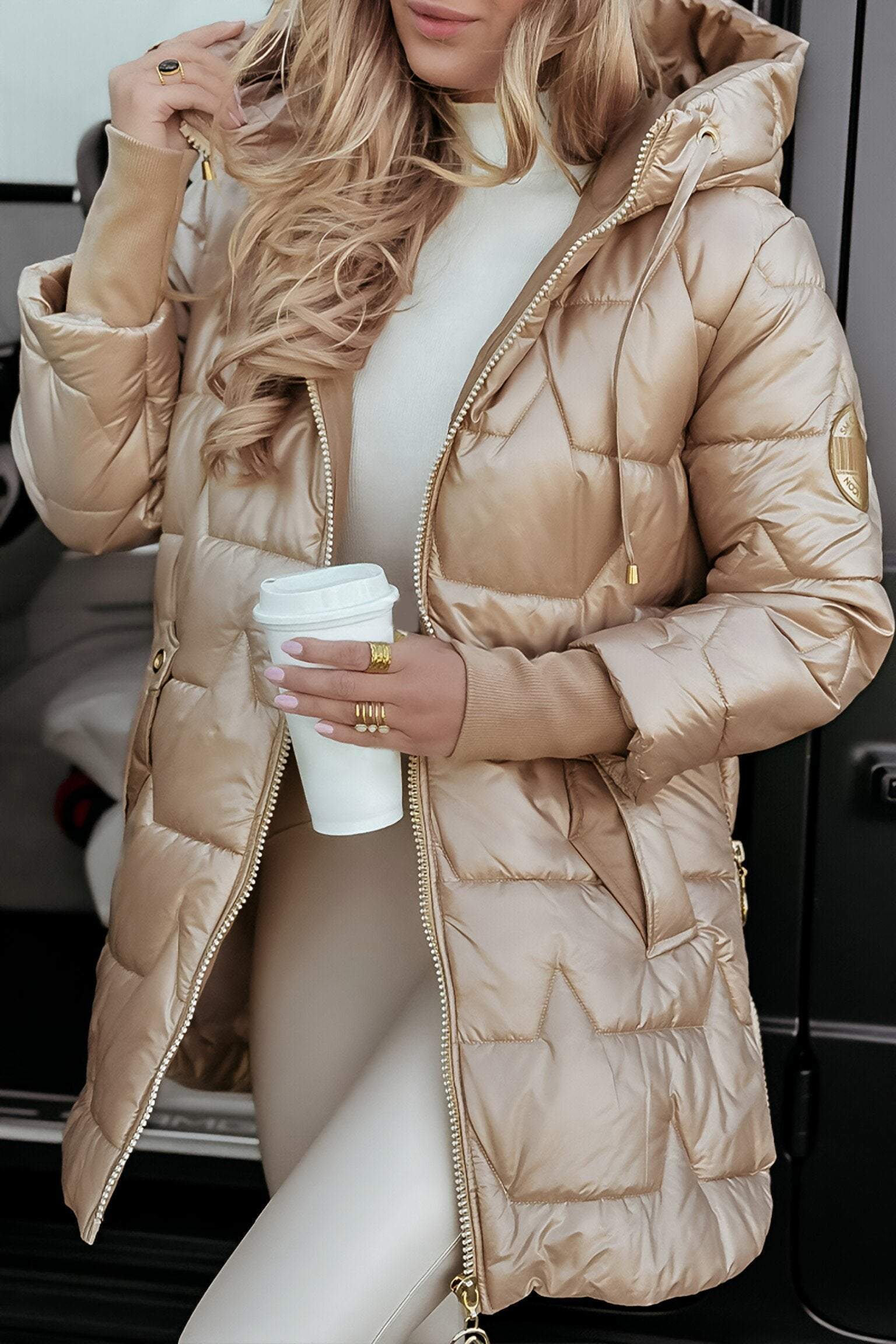 Femme en manteau matelassé doré, tenant un gobelet de café, devant une voiture. Mode hiver élégante, tendance, confort et style urbain.
