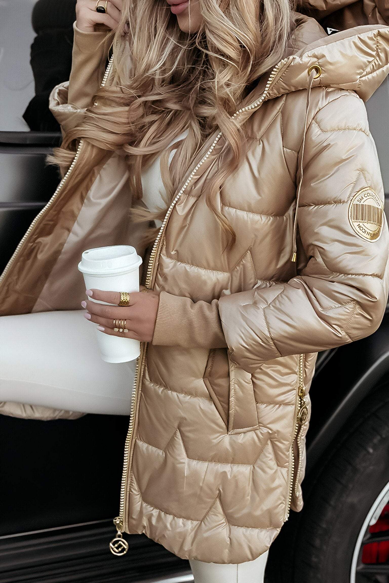 Femme en manteau doudoune beige tenant un gobelet de café devant une voiture noire. Mode hiver, style chic, tendance automne-hiver.