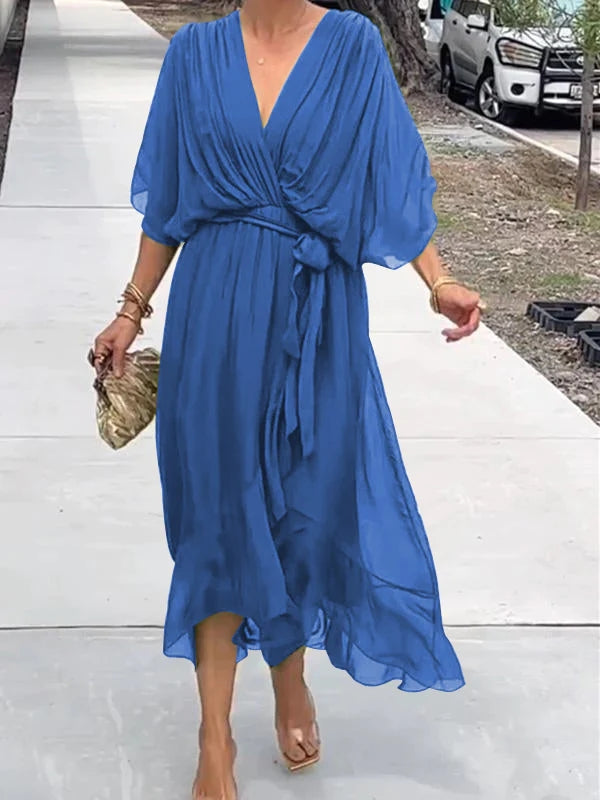 Robe bleue fluide élégante portée par une femme marchant sur un trottoir, accessoirisée d'un sac à main doré et de bracelets, style estival chic.