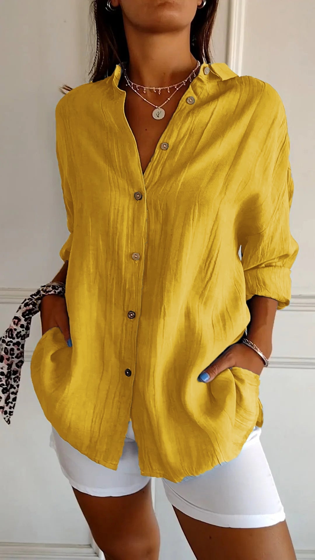 Femme portant une chemise jaune à manches longues, boutons en bois, col ouvert, avec un pantalon blanc. Style décontracté et moderne.