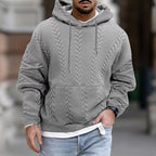 Homme portant un pull à capuche gris texturé, style décontracté, manches longues, sur fond urbain. Mode masculine automne-hiver tendance.