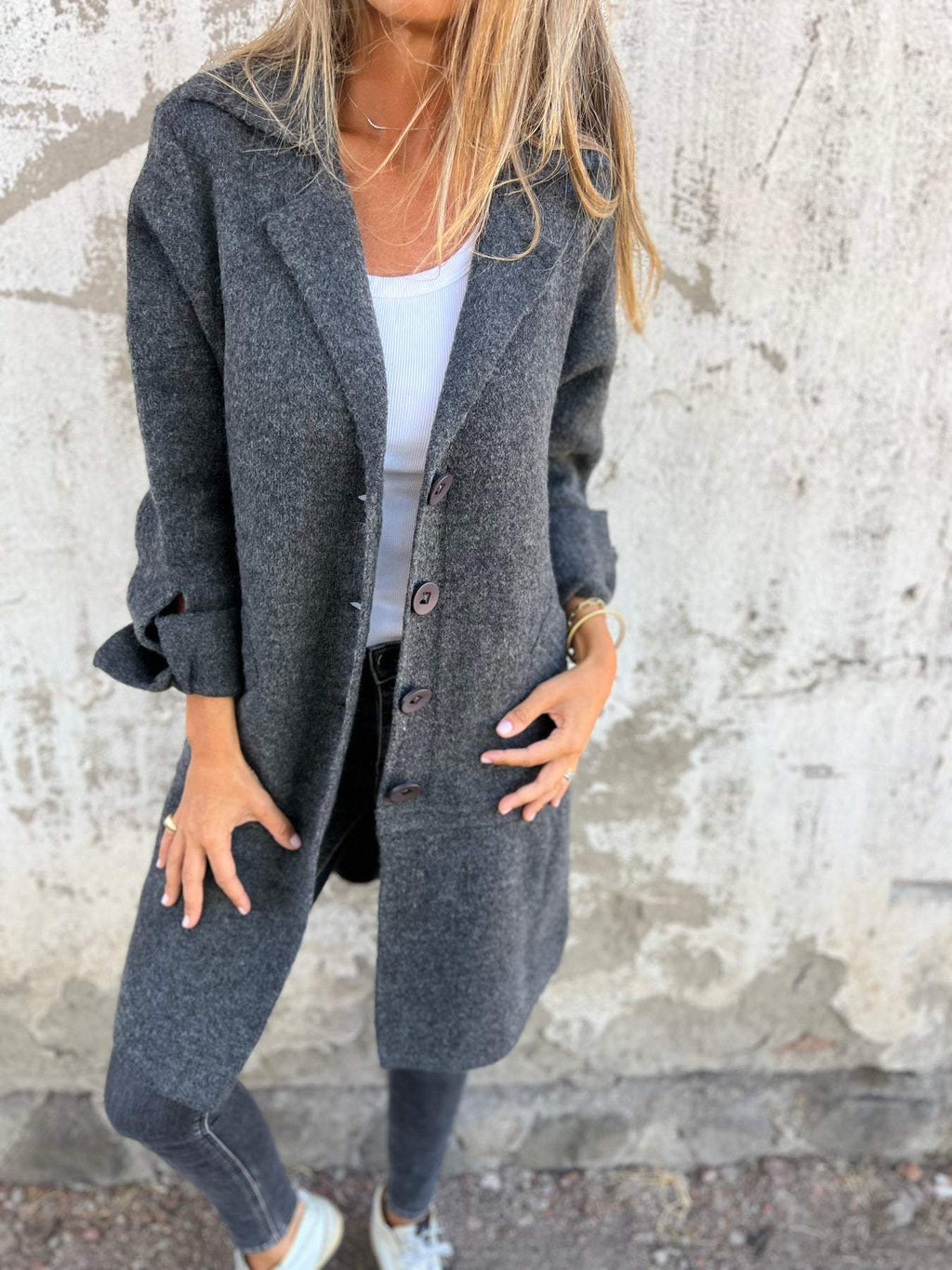 Femme portant un manteau gris en laine, chemise blanche et jeans, posant devant un mur texturé. Mode automne, style décontracté, tendance urbaine.