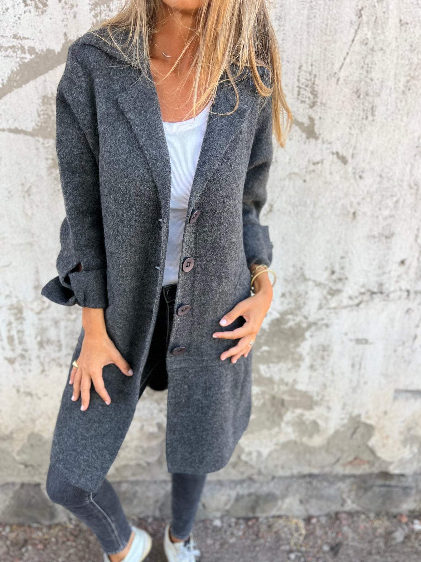 Femme portant un manteau gris en laine, chemise blanche et jeans, posant devant un mur texturé. Mode automne, style décontracté, tendance urbaine.