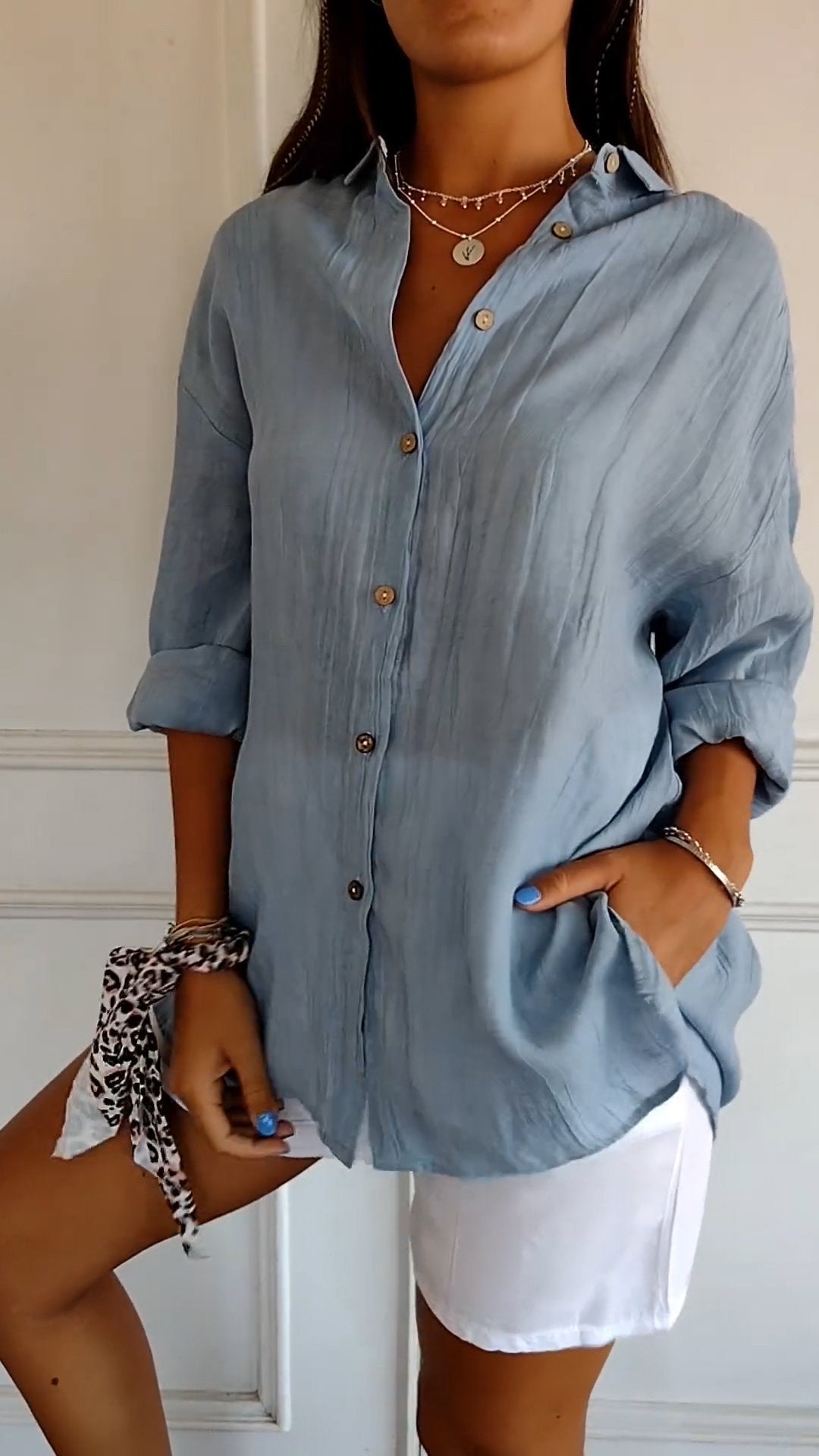 Femme portant une chemise bleue en lin avec boutons, manches retroussées, associée à un short blanc. Style décontracté, mode estivale tendance.