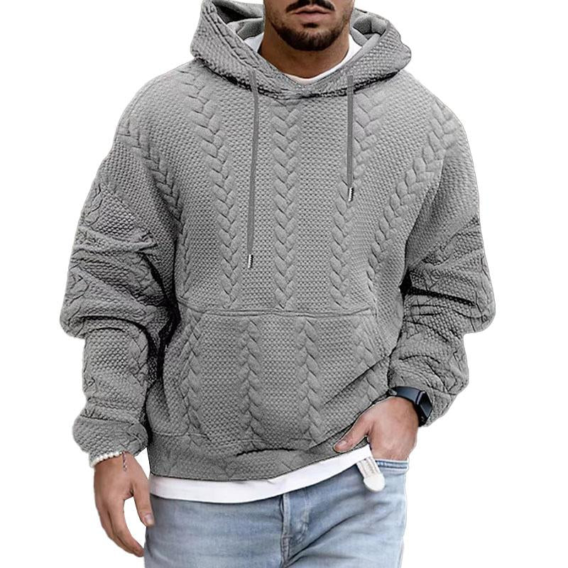 Homme portant un sweat à capuche gris texturé, style tricot torsadé, avec jeans décontracté. Mode masculine automne-hiver, look urbain tendance.
