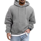 Homme portant un sweat à capuche gris texturé, style tricot torsadé, avec jeans décontracté. Mode masculine automne-hiver, look urbain tendance.