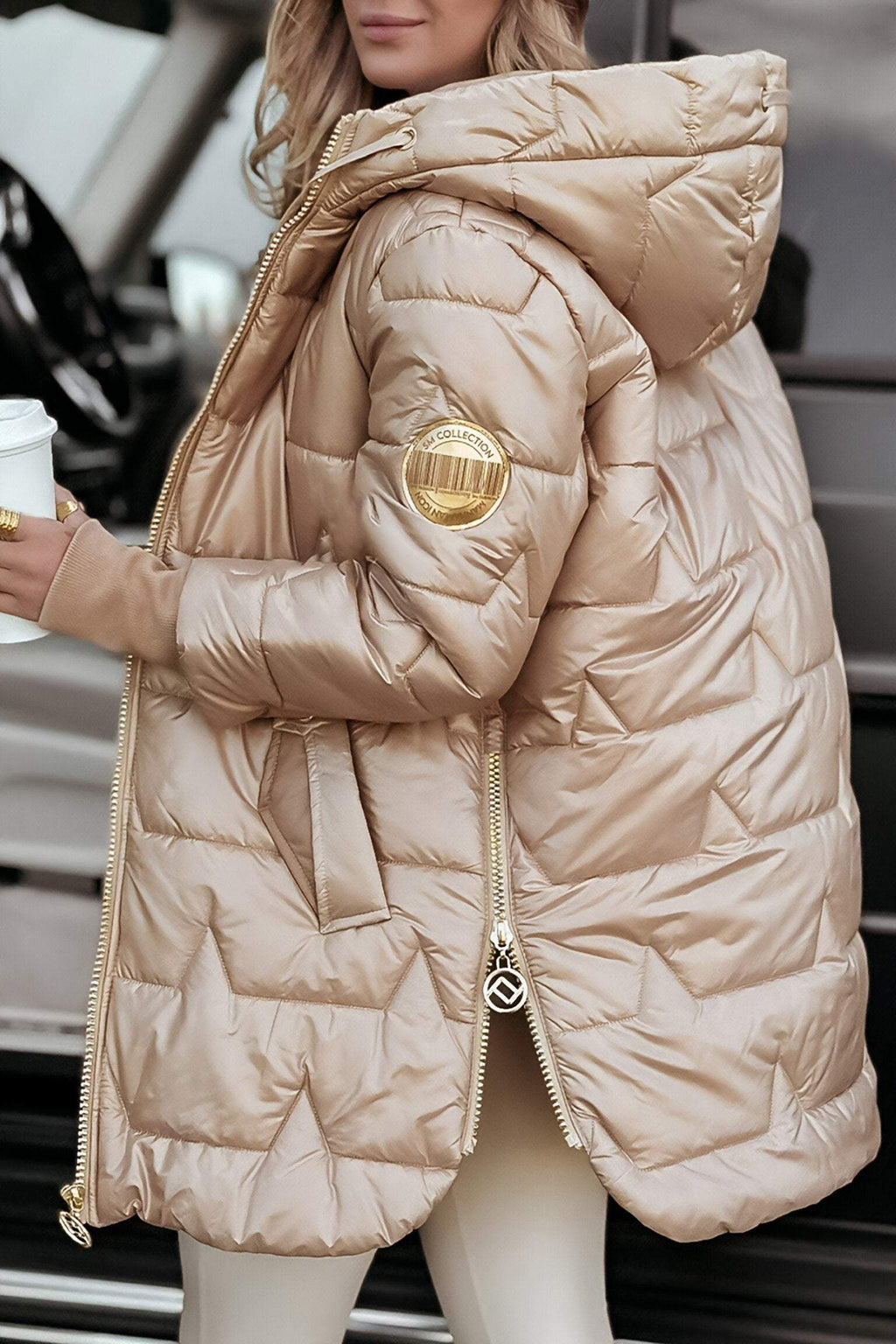 Femme portant une doudoune beige avec capuche, tenant un gobelet blanc. Veste matelassée élégante, idéale pour l'hiver. Mode féminine tendance.